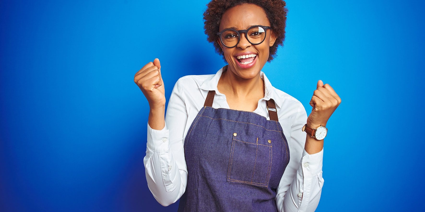 Young african american woman shop owner wearing business apron over blue background celebrating surprised and amazed for success with arms raised and open eyes. Winner concept.