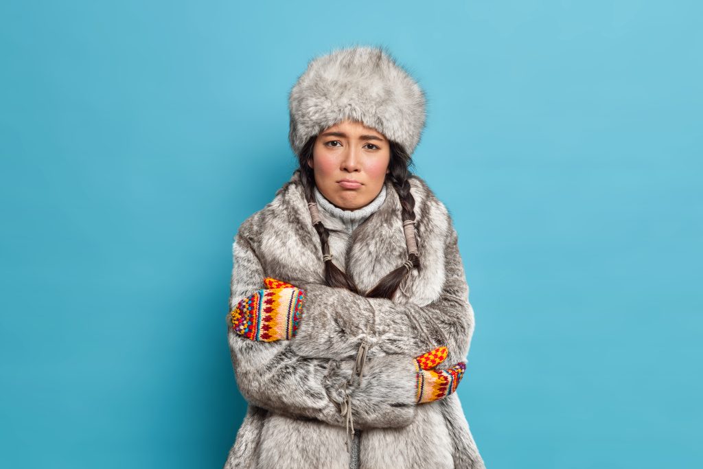 Unhappy scandinavian woman in fur hat and coat crosses hands and feels freezing trembles during severe frosty day wears winter outerwear. Yakut girl shivers from cold poses against blue background